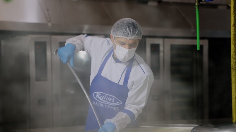 Kalori önlüğü, bone ve maskeyle kazanda yemek karıştıran aşçı
